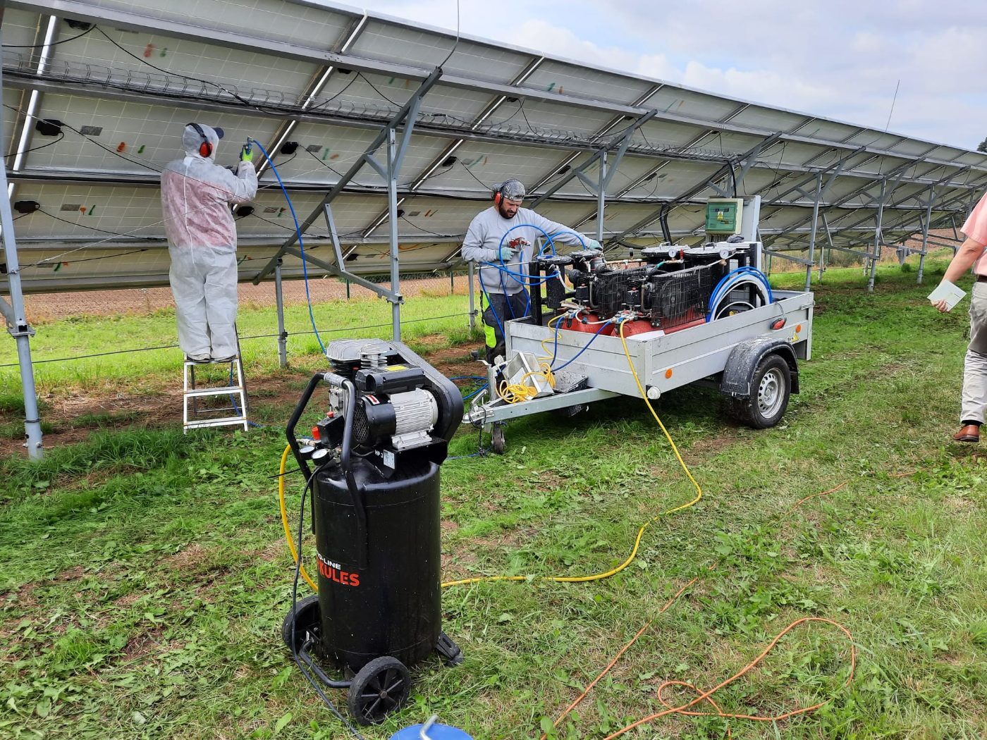 On-site renovation in progress — a technician in a white protective suit renovating panels from a ladder while a colleague works at the mobile silicone-gel rig, on a ground-mount array, photographed by Vladislav Poulek.
